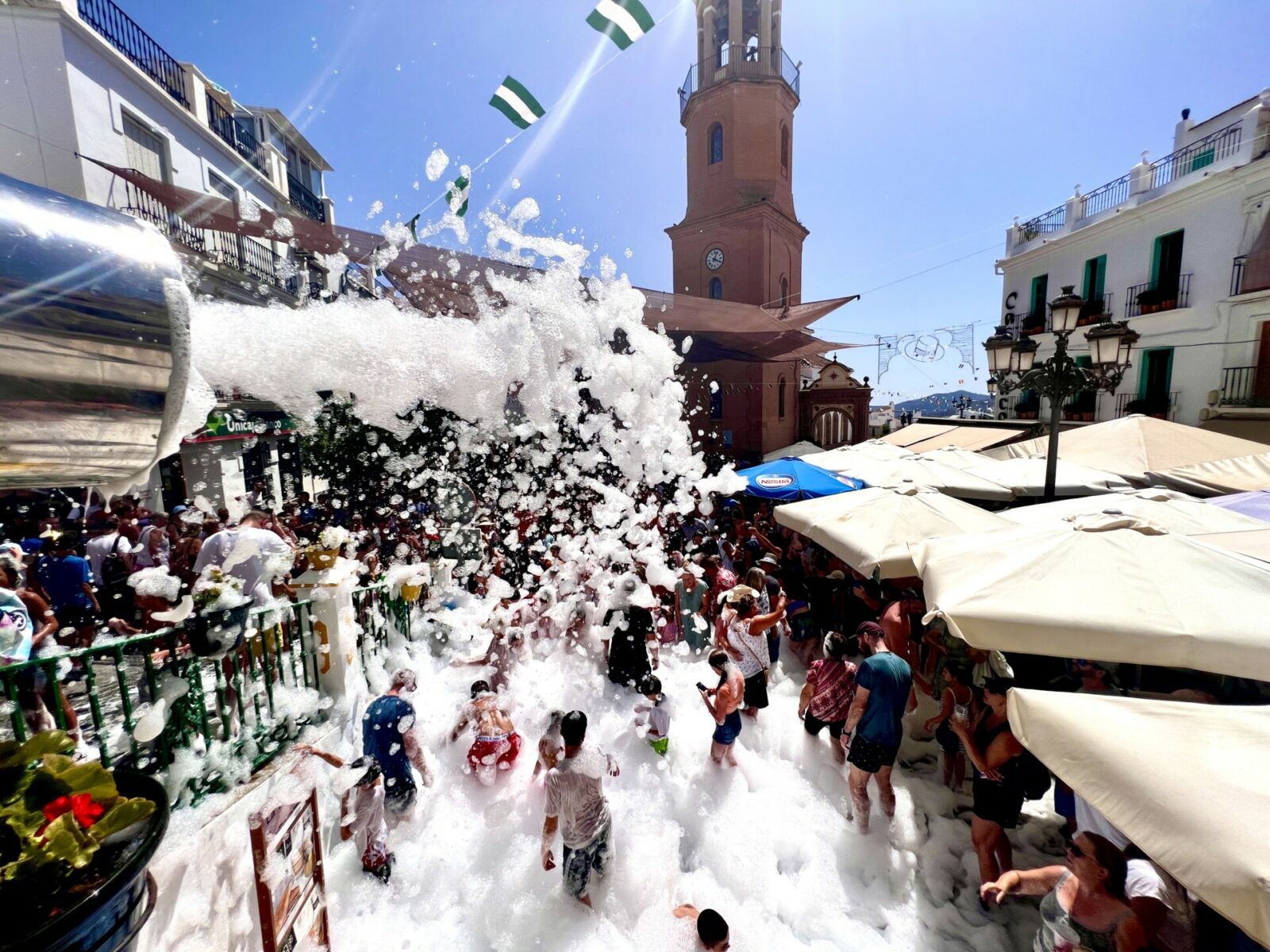 The image shows the church tower and the main square of Cómpeta, with white foam filling the square during the foam party of the Cómpeta Summer Feria.